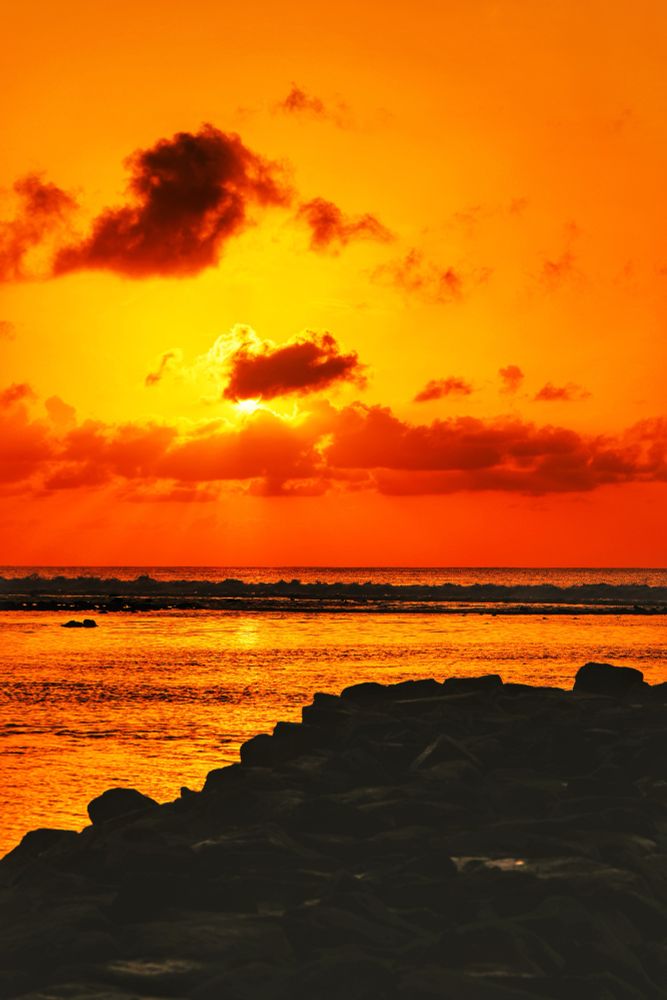 Hulhumalé, Maldives. Sunrise Golden Hour. Standing on the groyne at the end of Phase 1 next to the canal looking out to the sea and the rising sun. Most of the foreground are the rocks that make up the groyne. The rest is the lagoon lit up in sunlight and gold. Beyond it the reef in silhouette and the darker blue of the sea. From the horizon to the sun the sky and clouds are in tangerine. The sun is shielded behind a small cloud. The sky and clouds around it are in gold. Then slowly darkens to tangerine the further away from the sun it gets. 