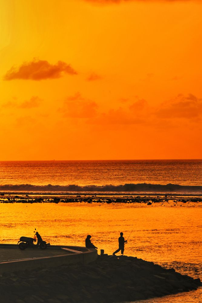 Hulhumalé, Maldives. Sunrise at the 2nd Phase sea wall. In the foreground is a parked scooter. Beyond that there are two people, one sitting on the sea wall boundary and another standing on the rocks and using a rod to fish in the lagoon. Everything is in silhouette. The lagoon is in gold. The reef is in low tide and the corals are showing above water and beyond that the waves crash. Both there too are in silhouette. The sea is simmering in gold and black. The clods in the sky are very faint and some nearly translucent. The sun is shining from the upper left of the frame and the color goes from gold to light tangerine. 