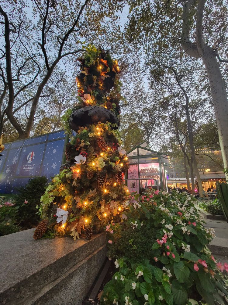 A decorative display of evergreens and twinkle lights at Bryant Park, alongside blooming pink and white flowers