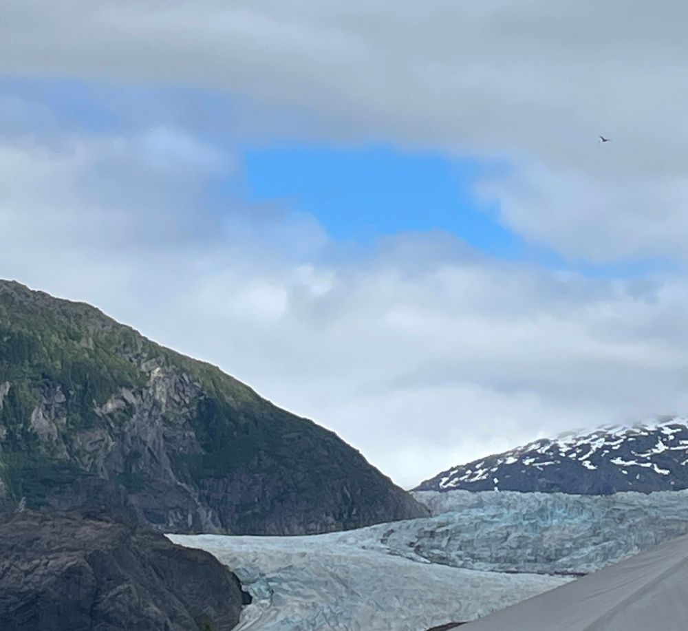 A spot of clear blue sky amid clouds atop snowy mountains, somebig green tufted rocks, and the Mendenhall Glacier showing off its white and blue flow. 