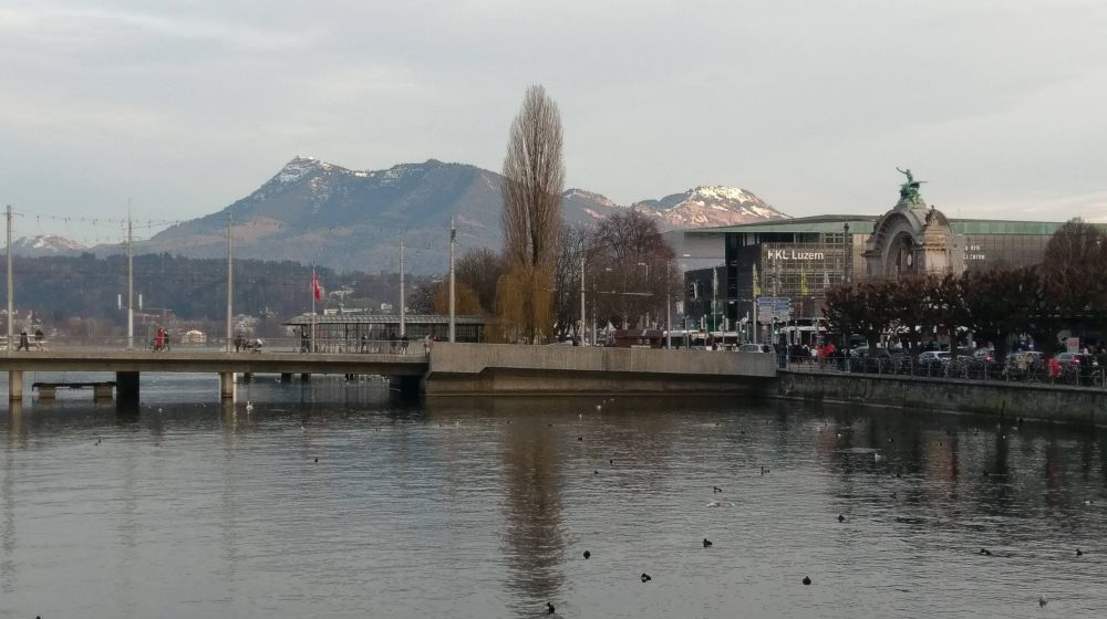 A view of some of the Alps in the distance from Lucerne, standing on the historic covered bridge so the foreground is water and then the other, modern bridge, and then beyond that the mountains.