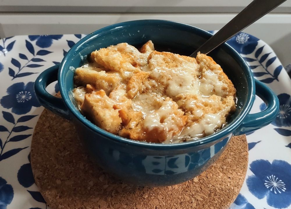 A crock of French onion soup. The top of the soup is broken pieces of crostini topped with melted cheese. It's all golden and toasty.