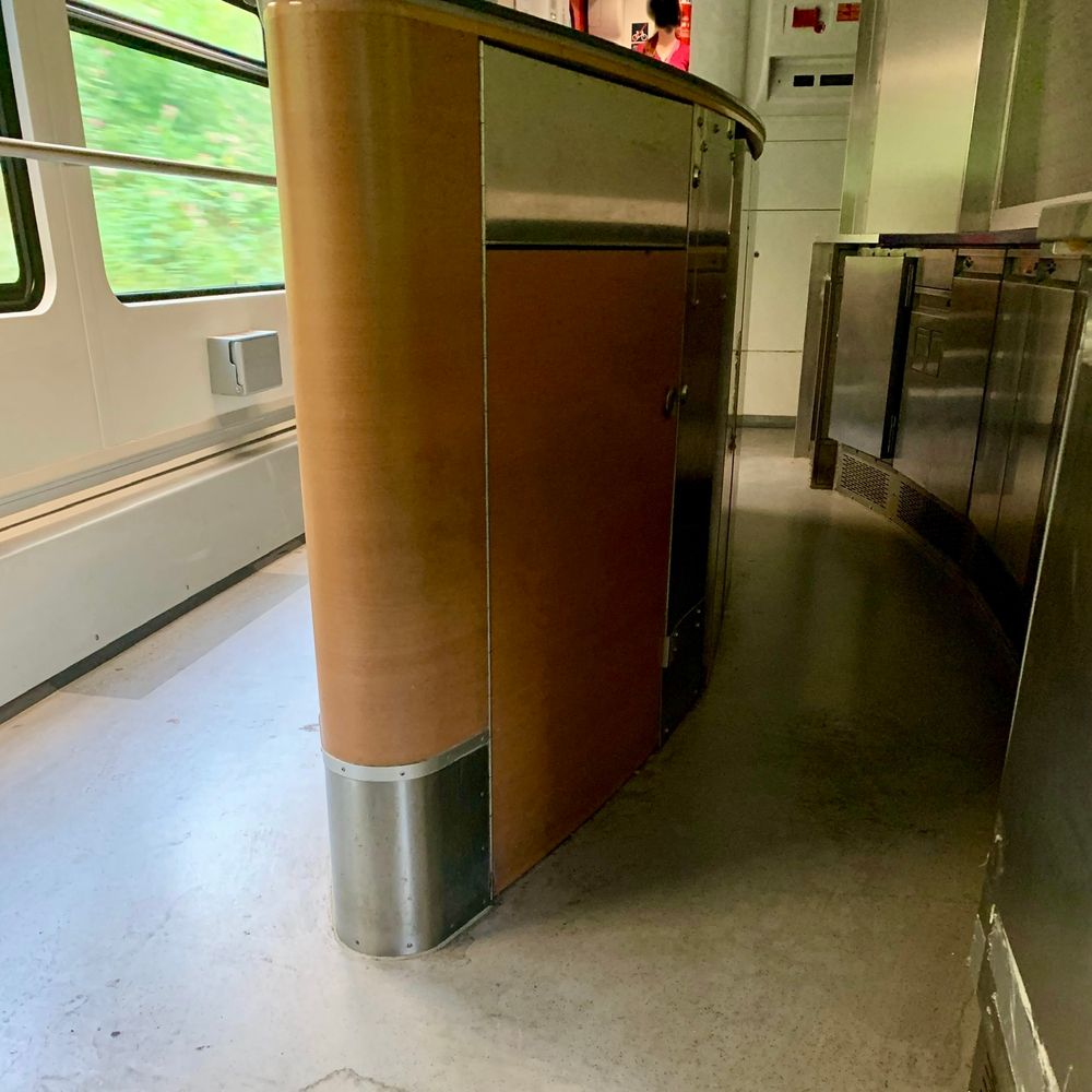 Eine alte Theke. Sie steht im Bahnabteil und ist oval und metallisch mit braun. Dahinter sieht man metallische Küchenschränke. 