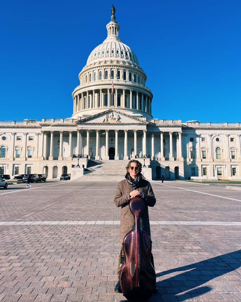 Photo of renowned cellist, composer and person living with Long COVID Joshua Roman standing in front of the U.S. Capitol with his cello, before meeting with key Congressional leaders to advocate for Long COVID research funding and patient support on December 12, 2024.