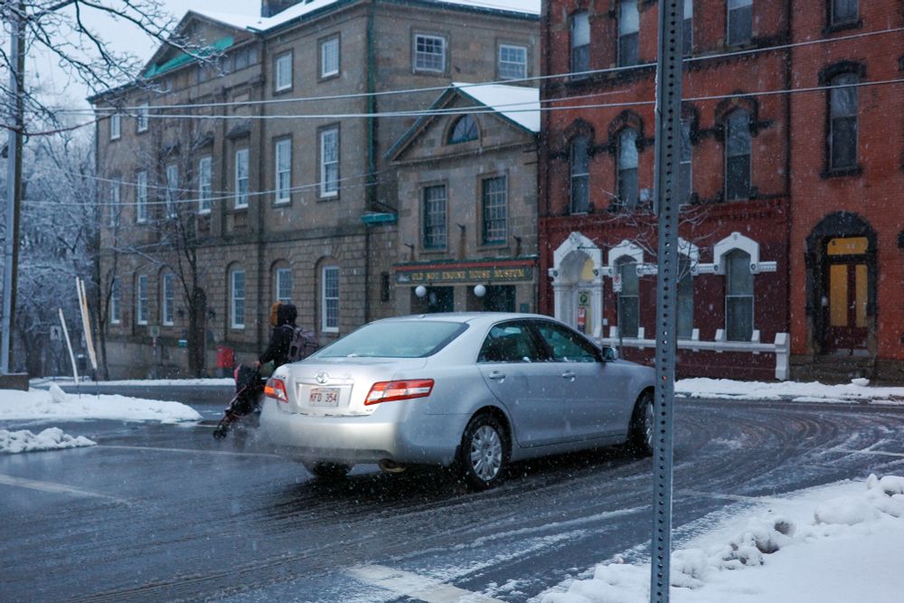 Silver car passing directly behind a mother crossing the street at a crosswalk pushing a stroller with snow and ice on the ground.