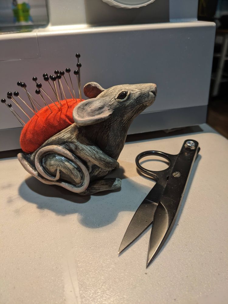 A realistic grey mouse sculpture with shiny black eyes and a back composed of a red pin cushion. There are 20 black-tipped silver pins piercing the cushion.  It sits on a grey table. It is pictured next to a grey sewing machine however only the bottom 1/5 of the machine is visible, and also next to a pair of metal snips, adding scale so that the mouse appears to be roughly 3 inches / 7 cm tall. 
