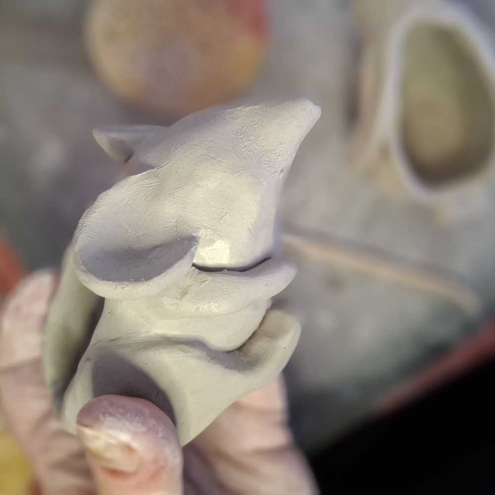 A wet white clay rodent-like sculpture held by three fingertips. The creature appears to be about 8-10 cm long.