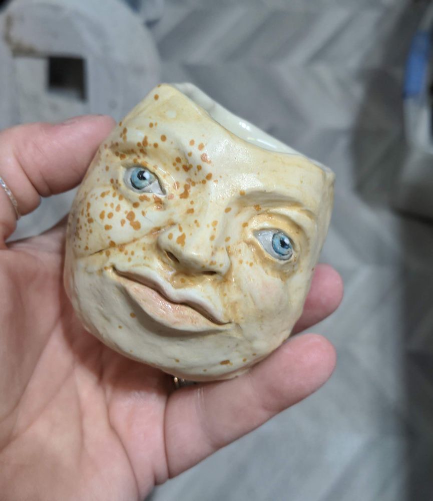Ceramic white clay head pot, held in a white woman's hand. It is painted with light beige skin, pale blue eyes that are very wide spread, pale pink wide lips, and brown random freckles across the left side of the pot. 