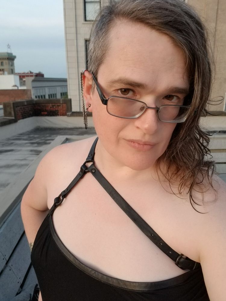 David, a white trans woman with glasses and a dark brown sidecut, this spring, wearing black dress with leather straps. She's smiling and looks far, far happier.
