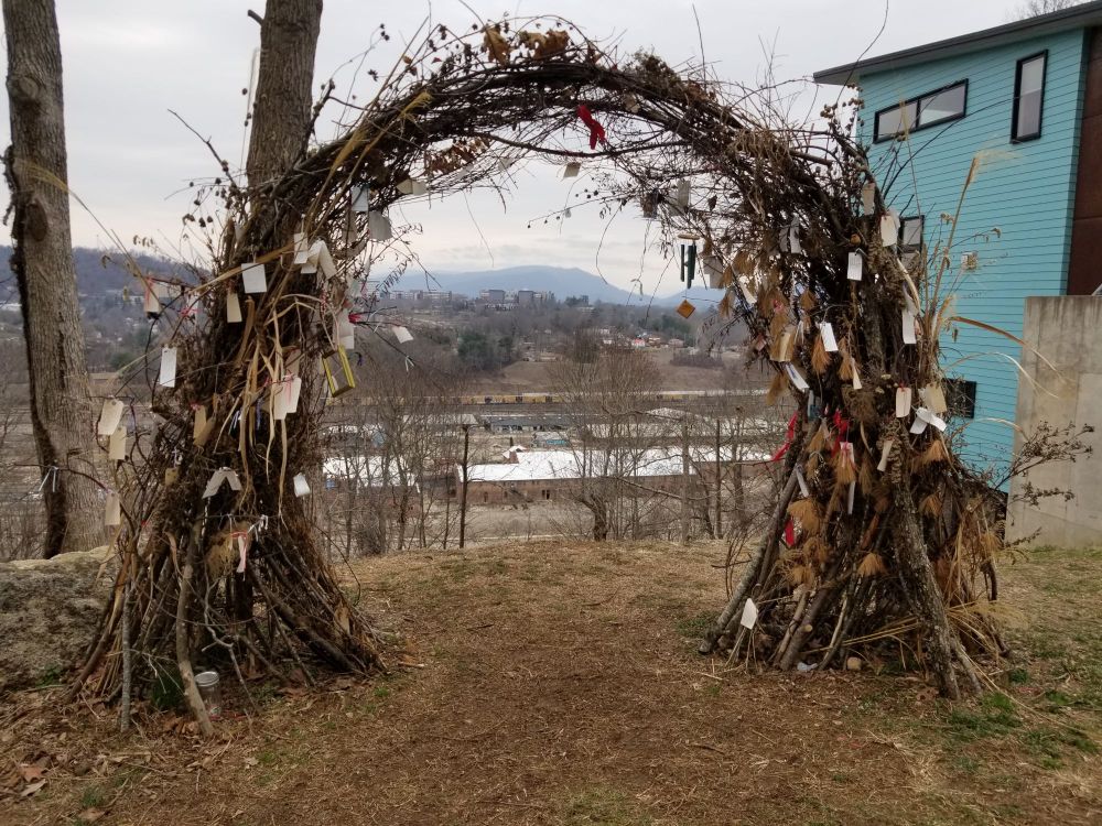 An arch of trees and branches with slips of paper hanging from it. A devastated area near a river is visible through it