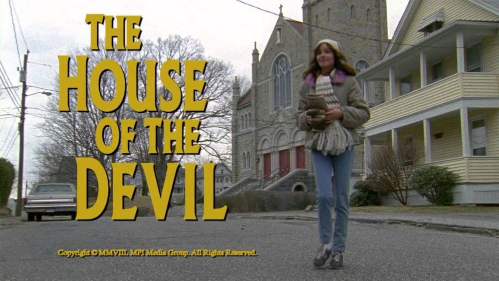 The title card to Ti West's THE HOUSE OF THE DEVIL with a young woman bundled up in the cold walking in street in front of a church and two story house 