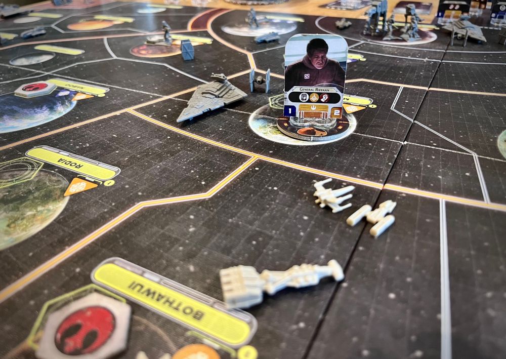 A close up of a dark board split into sections with orange and white lines. A standee token depicting a man looking down is surrounded by small plastic model ships. 