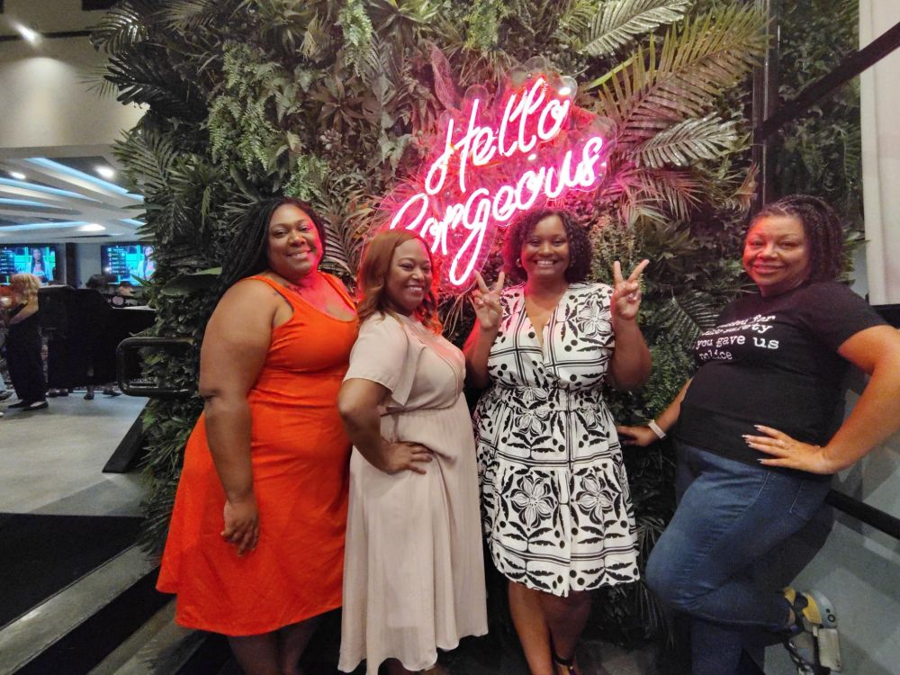 Photo of four Black Caucus leaders standing in front of a neon sign that reads "Hello Gorgeous" with a leaf background and smiling at the camera