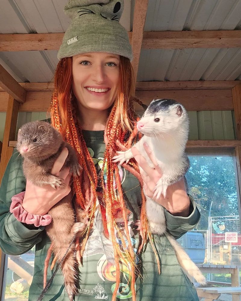The image shows Mikayla Raines smiling. She has long, bright red and orange braids wearing a green beanie with a simple black face design and a dark green sweatshirt that has colorful artwork and a small white logo reading "Uplift." She is standing in a sunlit wooden enclosure or shelter with a metal roof.

In each hand, Mikayla is holding an animal. In her left hand (right side of the image), she is holding "Scarlett", a dark brown mink with thick fur, a small pink nose, and dark eyes. In her right hand (left side of the image), she is holding "Atlas", a striking mink with a mostly white body and a black “cap” of fur covering the top of his head and neck. He has pink eyes and nose, and appears alert, facing toward the right. Sunlight is streaming in, casting warm highlights on all three of them. The atmosphere feels cheerful and cozy.