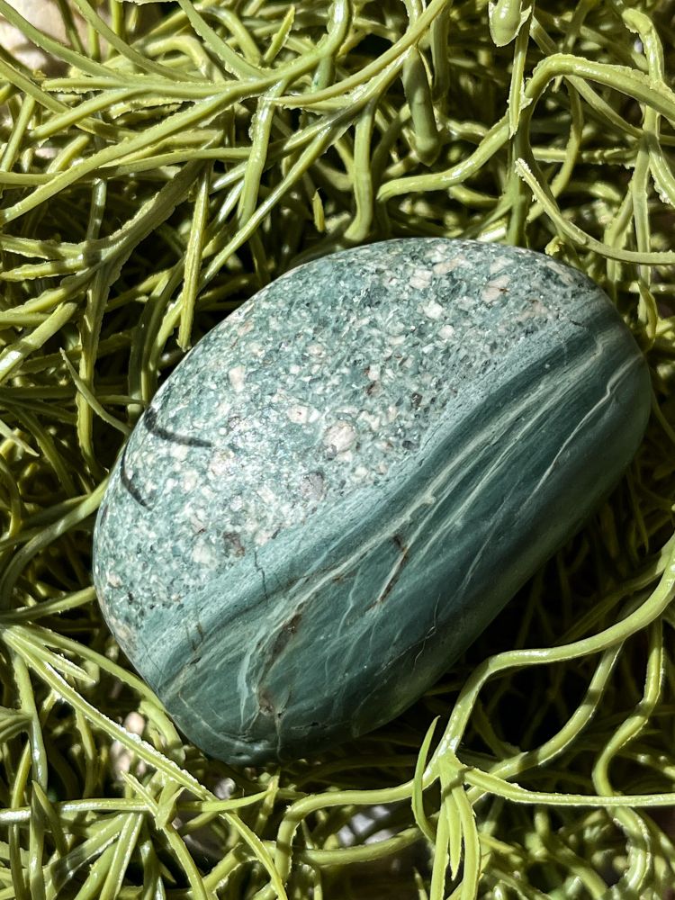 An oval shaped rock, half is a green and white speckled texture, and the other half is vertical stripes of various shades of green. 