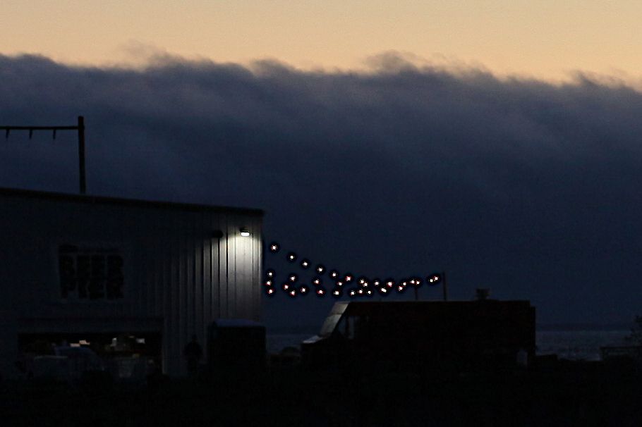 An extreme crop of the area by the warehouse's light in the full picture. You can make out the light shining on the windshied of a delivery van, the building's siding, and a dark window past some items in the yard. The clouds with the last light of the sun radiating past their tops is visible in the background.