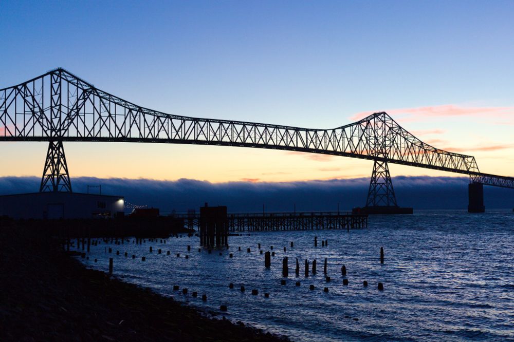 The Astoria-Megler bridge at dusk. It's an open iron construction, with the main deck suspended almost 200 feet off the water by a pair of tall towers. The Columbia's waters are a dark blue with some lighted areas across the waves. The dark remnants of old piers are jutting up in the foreground as the shoreline curves around to a warehouse and pier that mostly in shadows. You can see a s string of lights coming from it.
THe sun is nearly set behind a row of dark clouds on the horizon giving way to clear skies.