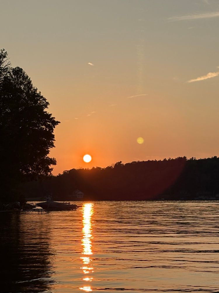 Sun setting over a hill on the side of a lake. The sun is reflected in the water, making it appear golden. 