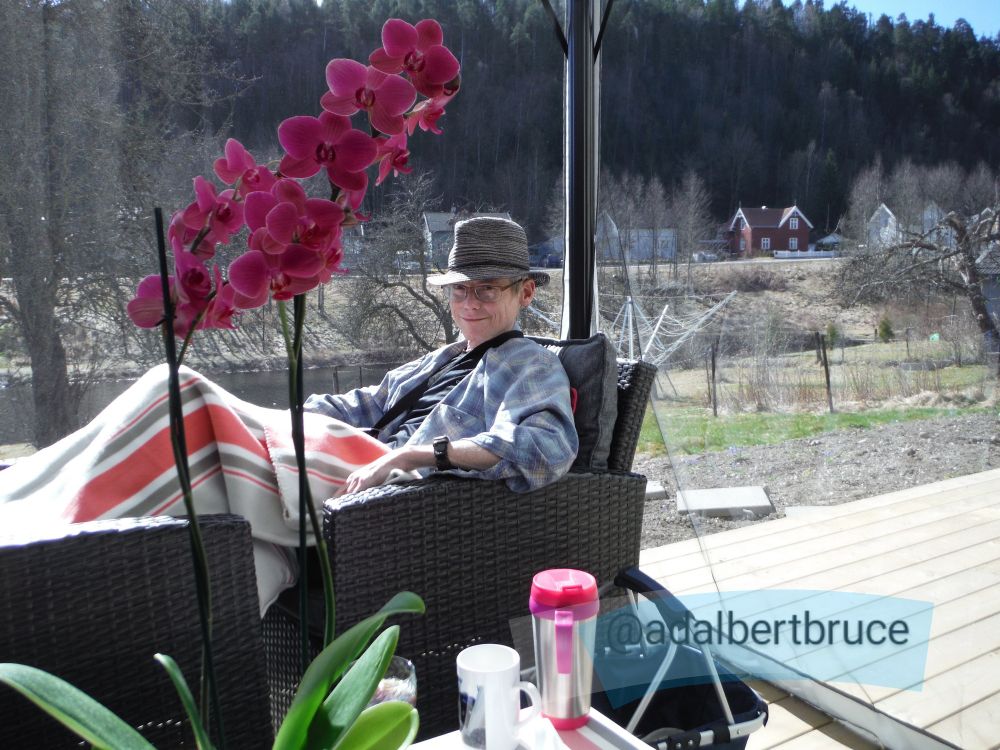 Artist Roman Scott, reclining, wearing a hat: With flowers in the foreground, and a forested ridge, and houses, behind. -JBF 