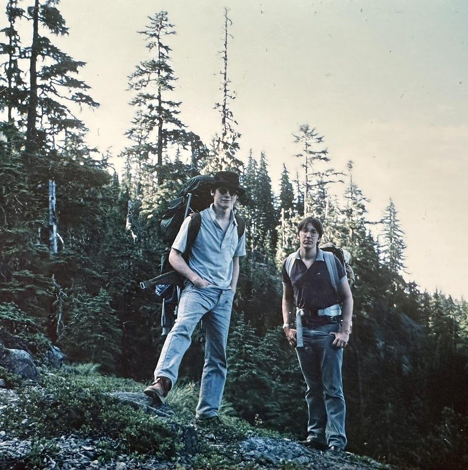 Roman Scott, artist, in black hat and photosensitive-dark glasses, and me, both with backpacks, atop a high rocky outcropping, with old-growth-appearing fir forests in the back; in Bull of the Woods wilderness, Oregon. Photo by Todd Mecklem. -JBF