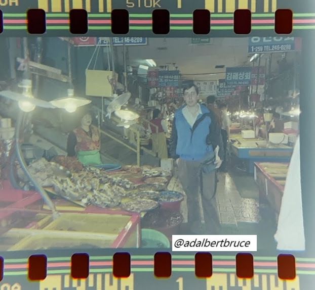 A photo of me, hands in pocket, with a blue windbreaker. I stood in Jagalchi Fish Market in South Korea, with bright lights hanging over seafood stands, with vendors. -JBF