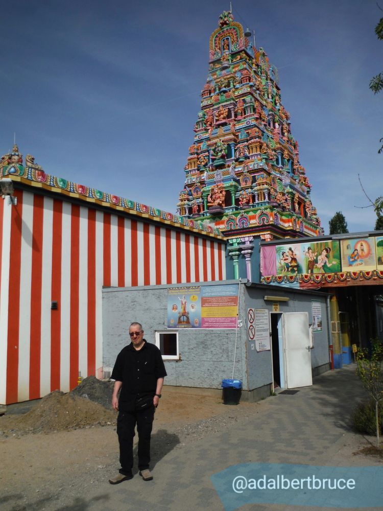 A photo of me, clad in black, standing before the striped and particolored backdrop of a Hindu temple in Germany. -JBF 