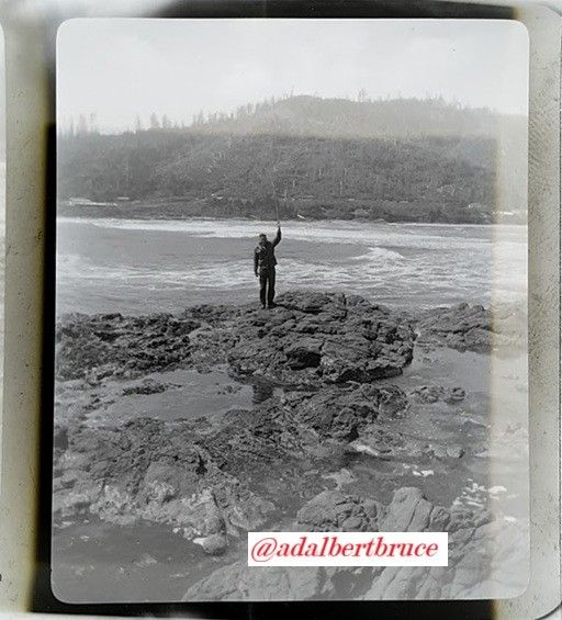Photo, possibly by, or of, Bruce W. Falk, showing (in black-and-white), a distant figure on a rocky shore, before surf, holding a stick, or fishing pole, vertically. Circa 1950. -JBF