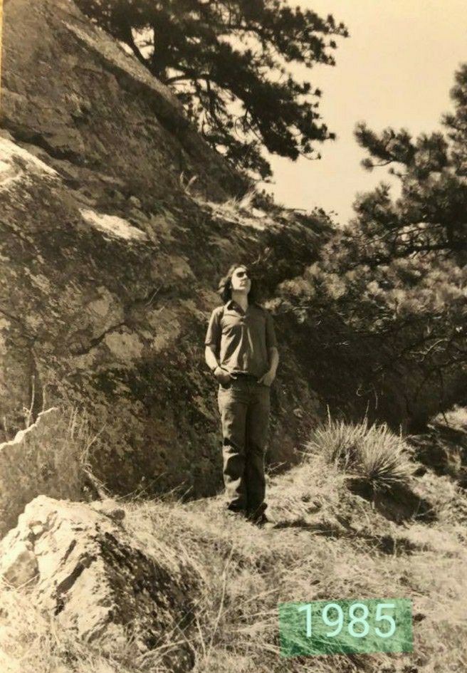 A photo of me, with dark lenses, and long hair, and hands in pockets. I was standing amidst pines, yuccas, and rocks, in the Boulder foothills. -JBF 