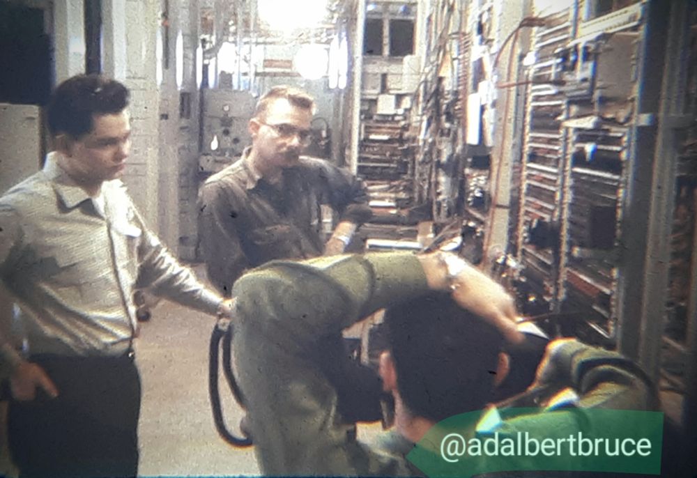 Slide photo by Bruce W. Falk. Three employees appeared, one in the foreground, back turned, arm over head. To the side was a wall of telecommunications equipment. -JBF 
