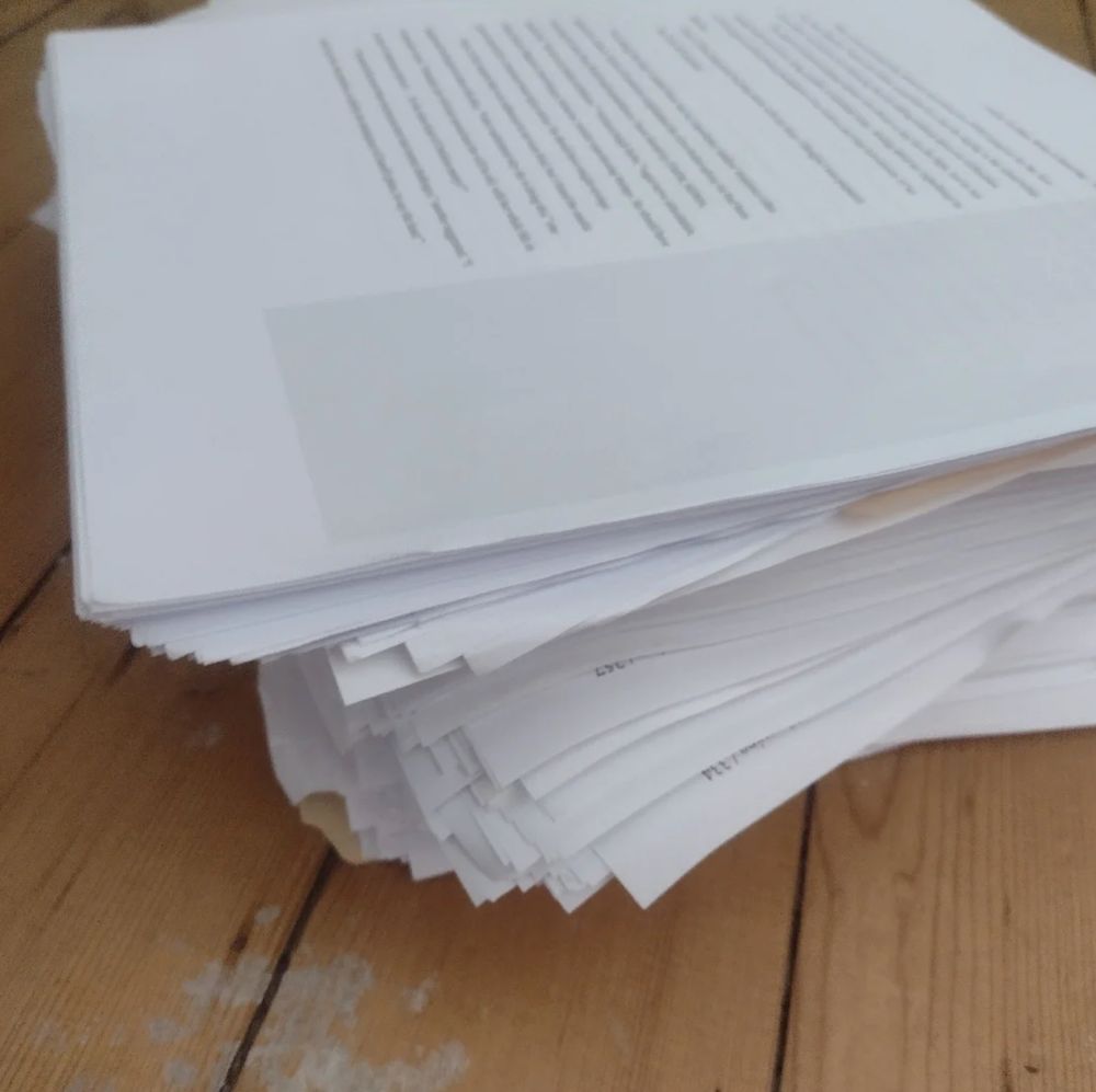 A large stack of drafts sits on a paint-spattered hardwood floor.