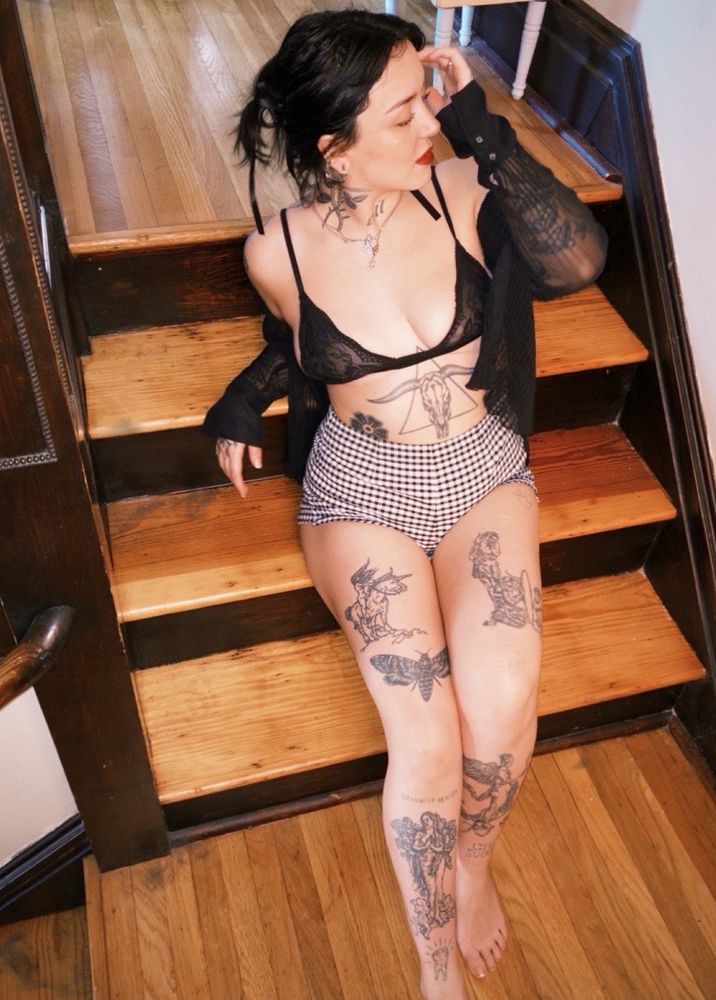 Billie is sitting on wooden stairs, wearing shorts and a small black bra and shrug, moving hair out of her face with all leg and stomach tattoos visible.