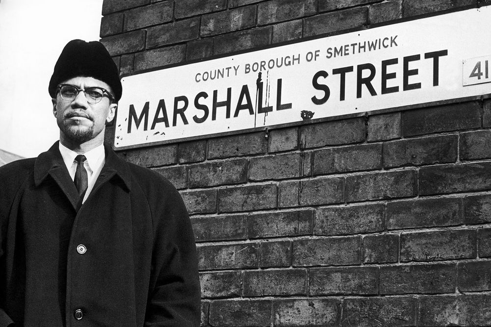 Black and white photo of Malcolm X standing in front of a brick wall and a street sign saying County Borough Of Smethwick, Marshall Street.