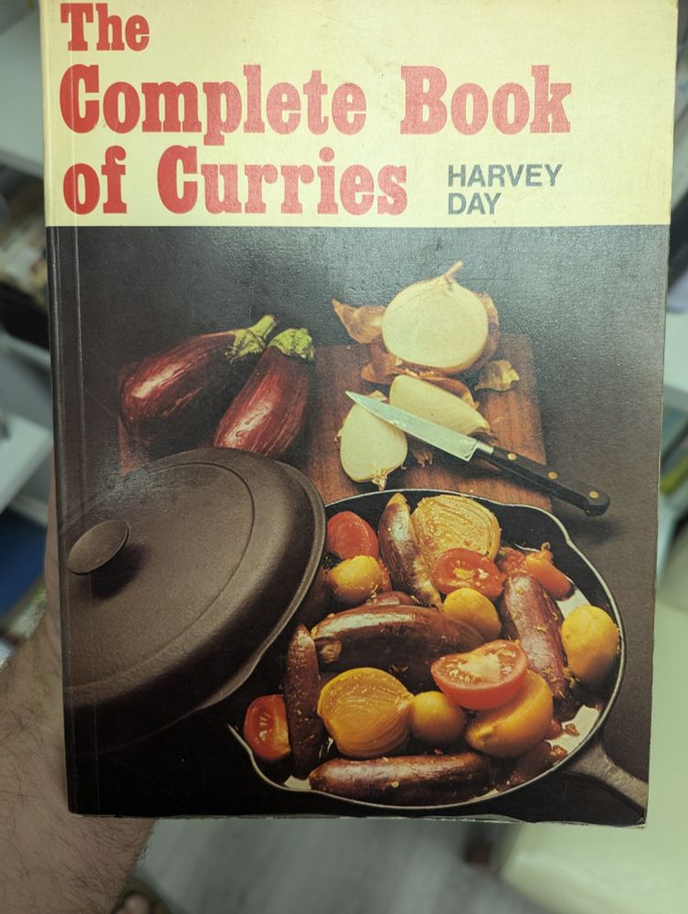 Cover of a cookery book entitled The Complete Book Of Curries by Harvey Day. The cover has a photograph of shallots, onions, tomatoes and aubergines in a cast-iron pan (although the shallots look more like sausages)