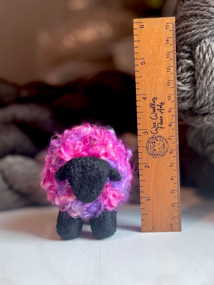 A handmade wool sheep with black legs and head and pink/purple body facing the camera. There is a wooden ruler to the right of it showing the sheep is just over 3 inches tall. There are skeins of grey yarn in the background, out of focus.