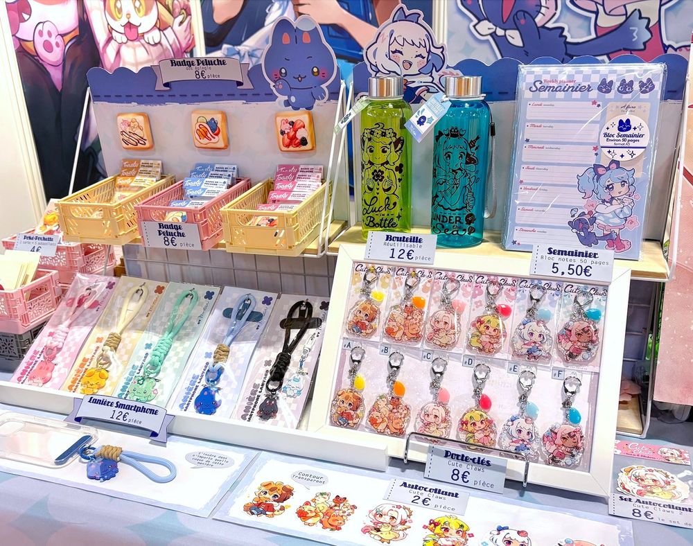 A close-up of an artist alley booth, showing original characters merchandise. We can see two bottles, keychains, phone lanyards and plush badges in a cute style.
