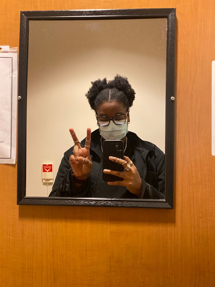 A mirror selfie of a Black person wearing glasses, a face mask, and a black jacket. Their hair is styled in two puffed sections, and they are holding a phone with one hand while making a peace sign with the other. Their fingers have several silver ring splints on them. The mirror is framed in black and mounted on a wooden wall, with part of a fire alarm and a posted document visible in the background.