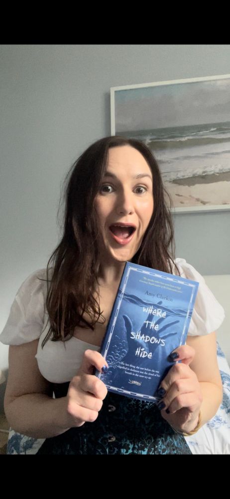 Amy, a white woman with long brown hair, is holding a blue book with an eerie hand reaching through water on the cover. The book is called Where The Shadows Hide and Amy’s expression is wide eyed excitement. 
