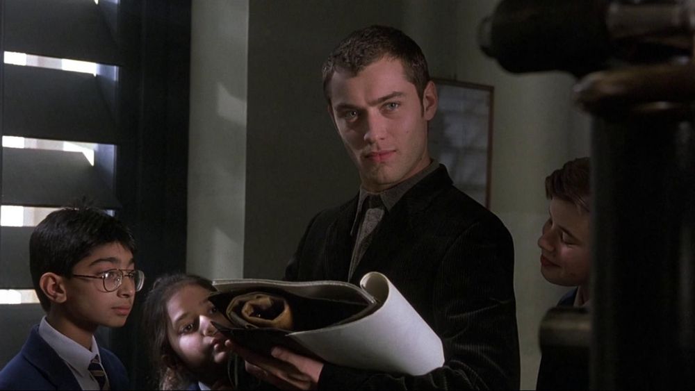 Jude Law regardant presque vers la caméra et entouré de trois enfants visiblement très intéressés par le carnet de dessin qu'il tient dans ses mains. Image tirée du film La Sagesse des Crocodiles