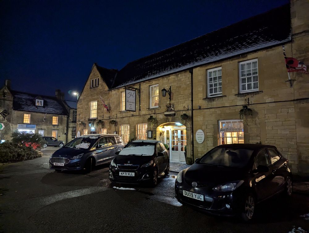 An older world coaching inn at night atmospherically lit. 3 cars are parked at the front, one with snow on its roof. 