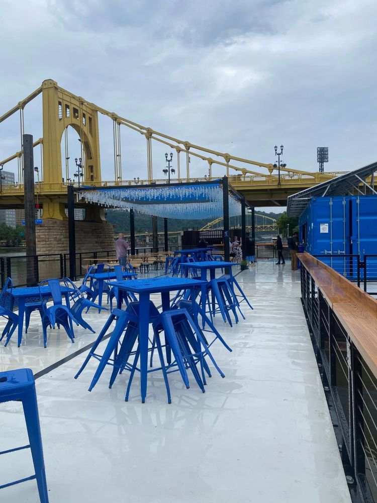a floating dock with blue tables and stools in front of a yellow bridge