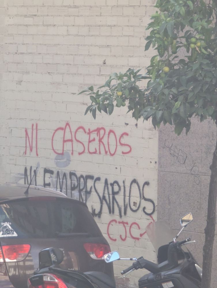 Pintada sobre un muro. Está parcialmente tapada por un coche y vista de lado, pues la foto se hizo desde un autobús en marcha. Dice: NI CASEROS NI EMPRESARIOS.