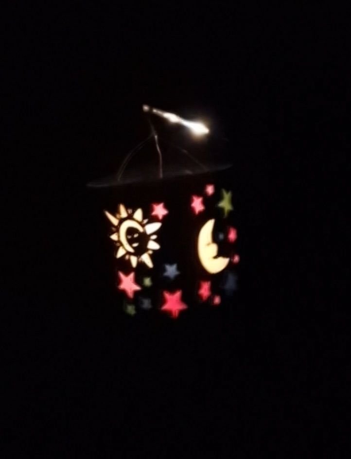 Eine viereckige Laterne,  beleuchtet in der Nacht. Man sieht eine gelbe Sonne, einen gelben Mond und bunte Sterne.