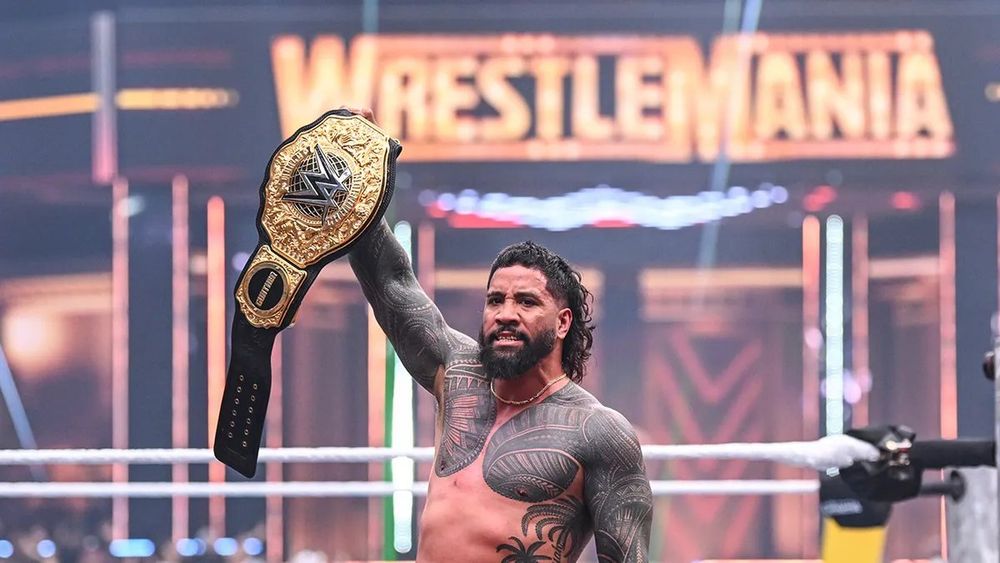 Jey Uso holding up the World Heavyweight Championship with the WrestleMania banner behind him