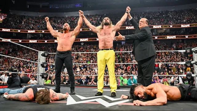 Bron Breakker and Seth Rollins holding both arms up high with Paul Heyman pointing to them as CM Punk and Roman Reigns are laid out on the ads-infested WWE canvas.