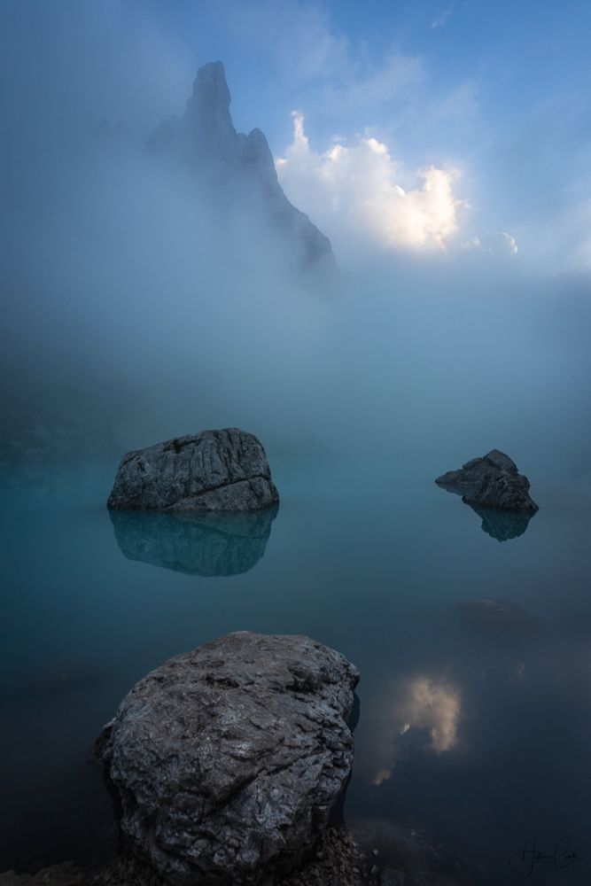 Three round rocks emerging from a turquoise blue foggy mountain lake with a rocky mountain peak appearing in the fog in the background in the left corner. In the right corner, the blue sky bathed in the late afternoon light and underlined with a puffy white cloud. All elements are relected in the lake.