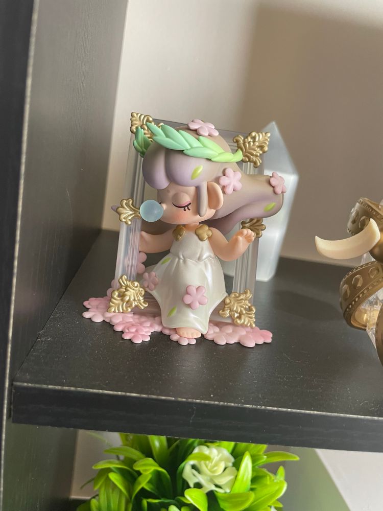 a cute museum figure of a girl with a bubble coming out of her face her eyes are closed, shes coming out of a painting frame. loonas lights stick is to the right.