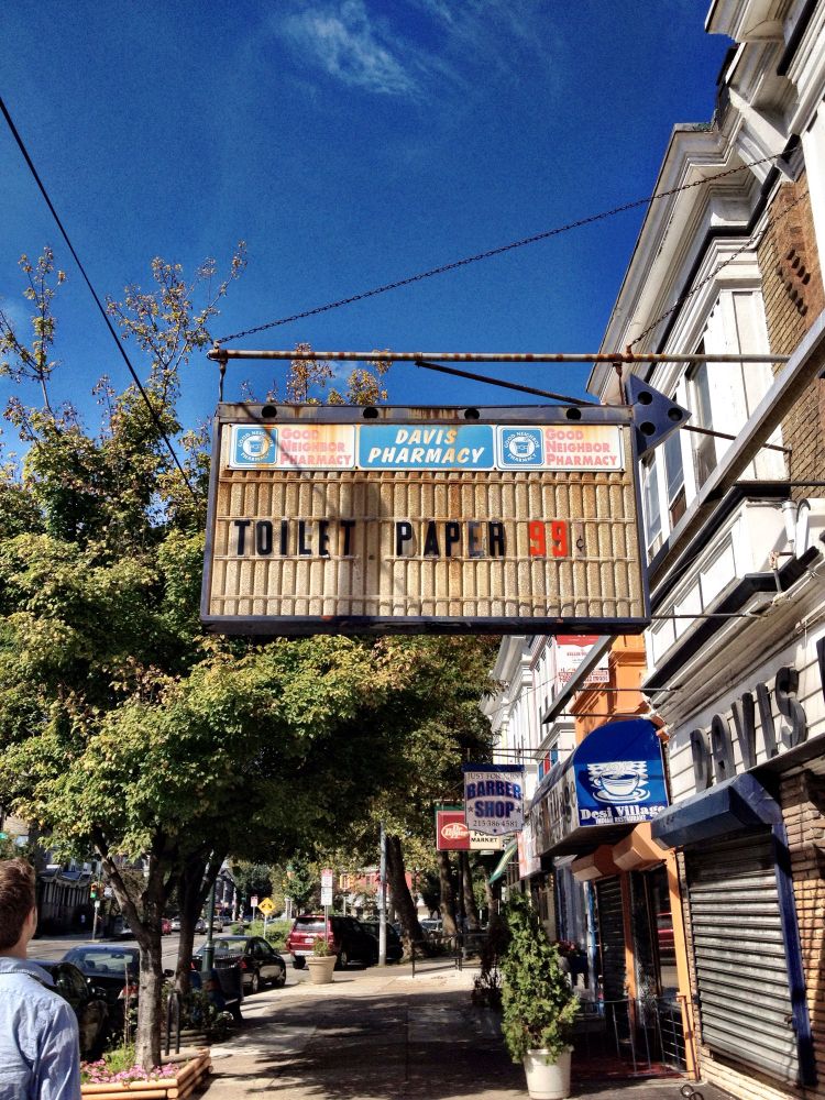 A street in Philly on a bright sunny day with a old Davis Pharmacy sign overhead, with hand-paced sign letters reading: TOILET PAPER 99 