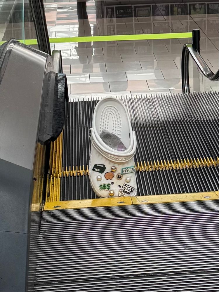 A Croc shoe sits wedged, toe in, heel up, in an escalator at the mall on a Sunday afternoon. No people are on the escalator and no person is with the now abandoned, escalator-decommissioning Croc. The white Croc has various Croc accessories, all money themed. “Lucky You!” “Money Maker” “$$$” and images of a piggybank, wallet and faux pearl and gold further adorn the shoe. The bling may have saved a toe. We can only speculate.