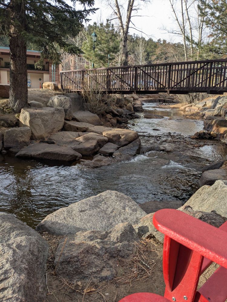 The edge of a trio of red Adirondack chairs along a gently babbling river framed by pine trees in Estes Park, CO. A bridge in the background, and coffee and book in hand, but not in the picture 