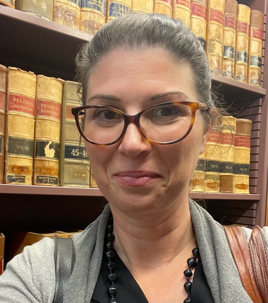 A middle aged woman who needs to push her glasses up and quit wearing her hair in a bun, standing in front of a bookcase of really old law books that she wants to spend hours reading but can’t because adulting is the worst.  